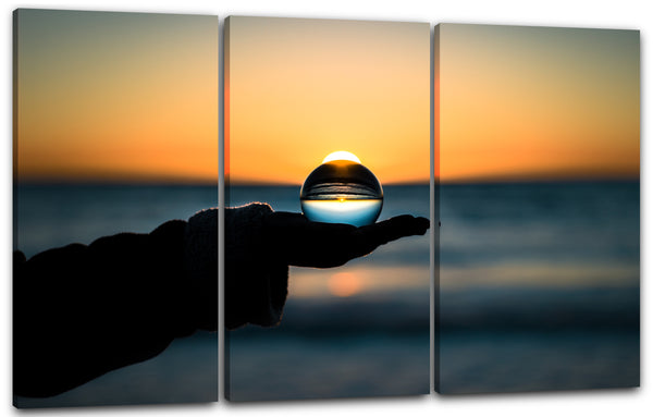 Leinwandbild Landschaftsbilder Glaskugel in Hand vor Meer und Sonnenuntergang