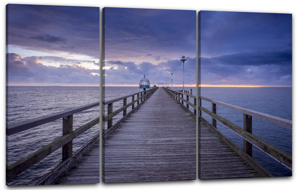 Leinwandbild Holzsteg am Meer mit Geländer und Laternen vor Himmel in violett